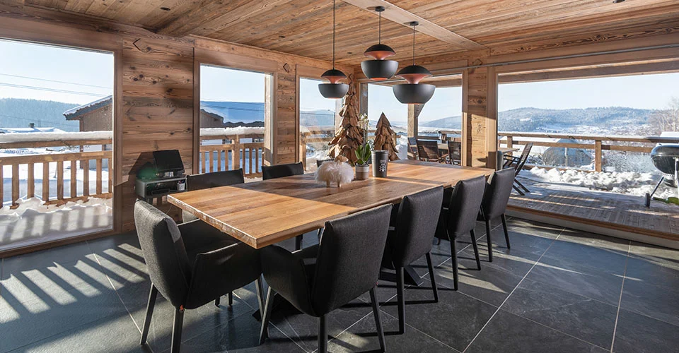 Interior view of a Decobois wooden chalet: warm wood ceilings, dining table, snowy Alps outside.